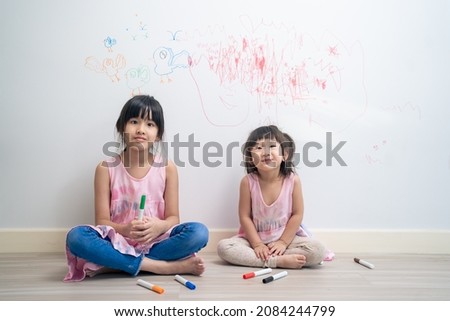 Asian young sibling kid girl enjoy paint on white wall in living room. Little adorable children having fun drawing and coloring art picture with hapiness enjoy creativity at home and looking at camera