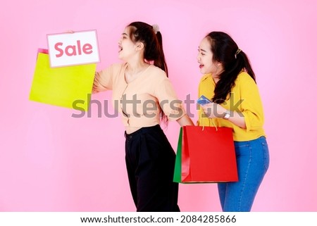 Studio shot of two Asian young happy cheerful female shopaholic shopper in casual outfit smile walk holding sale discount banner placard sign colorful shopping bags and credit card on pink background.