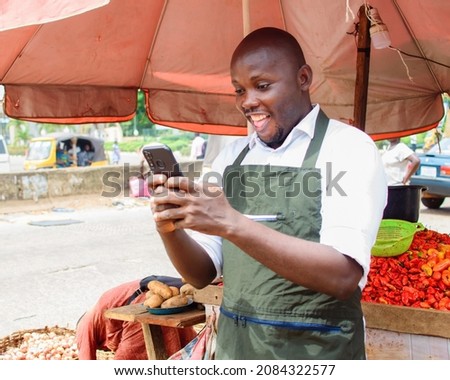 A happy African male trader with a smart phone, standing beside his stall of tomatoes and pepper in a market place Royalty-Free Stock Photo #2084322577