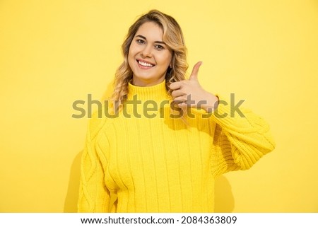 Young caucasian cute happy blonde woman in sweater isolated on yellow background, giving thumbs up gesture and smiling with teeth looking at camera. Possitive emotion. Studio shot