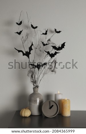 Alarm clock, burning candles and vase with paper bats  on table near white wall. Halloween decor