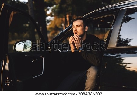 Driver man sitting in the black car on nature and look shocked. Ordinary slender european male model with beard holding hands on cheeks with opened mouth and shocked expression, being shock and scare. Royalty-Free Stock Photo #2084366302