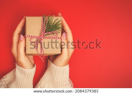 A Christmas gift in kraft paper, decorated with a coniferous branch in women's hands on a bright red background.