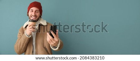 Young man in winter clothes and with cup of coffee taking selfie on color background with space for text