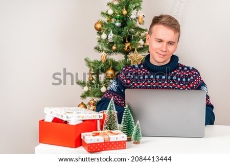  A young man dressed in a sweater using a laptop at his desk next to a Christmas tree and a cozy New Year atmosphere. The concept of online shopping and communication