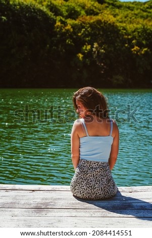  A charming young woman in a skirt is sitting on a wooden bridge against the background of nature with a lake in the forest. Travel, freedom, the concept of an active lifestyle.