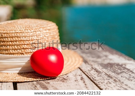 A red heart and a straw hat are lying on a wooden pier on the lake. Beautiful summer concept of a serene holiday.