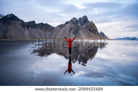 Iceland landscape photo of brave girl who proudly standing with his arms raised in front Vestrahorn mountaine on Stokksnes cape in Iceland.  Amazing Iceland nature seascape, Water reflection