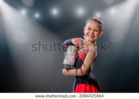 Portrait of a happy little skater with skates in her hands on the ice arena, dark background