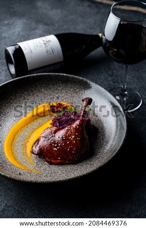 Duck thigh and carrot puree on a wide dish, bottle and red wine glass on grey background, side view