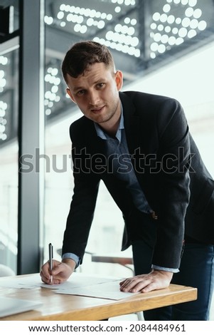 Confident young business man signing documents in a modern office with window in background. Pen in hand, papers on the wooden desk, futuristic background.