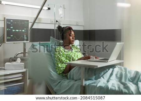 African american sick patient standing in bed during medical appointment watching online videos on laptop computer. Young woman recovering after disease surgery in hospital ward. Medicine service
