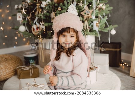 little girl in a winter fur hat sits on the floor near the Christmas tree with gifts.