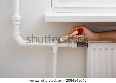Young adult man hands using brush and painting heating pipe white color. Closeup. Repair work of home. Royalty-Free Stock Photo #2084563042