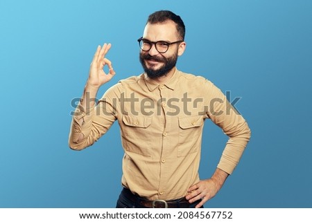 Positive adult worker male wearing eyeglasses and beige shirt showing OK gesture and looking at camera with excited expression against blue background
