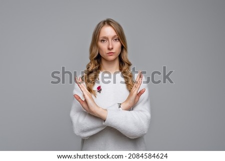 Serious blonde young woman wears white sweater showing stop gesture with crossed hands, trying to defend herself as if saying: Stay away from me. Stay home. Social distancing. New normal behavior