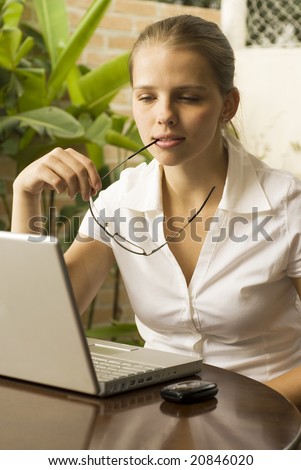 Thoughtful woman at a computer with her glasses between her lips. Vertically framed photo.