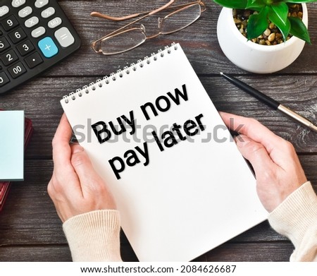 Hands hold notebook with text BUY NOW PAY LATER  near glasses, calculator, green plant with a pen on a wooden table. Flat lay, close up