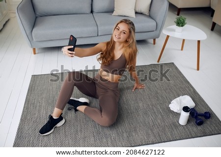 A sportive girl is doing selfie while looking at you. She has dumbbells and a bottle of water near her. She has a happy face. There is a gray sofa behind her.