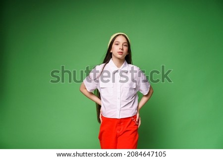 Cute pretty smiling caucasian teenage girl in hat and white shirt and red pants standing isolated on green background. Looking at camera, close up portrait posing for casting