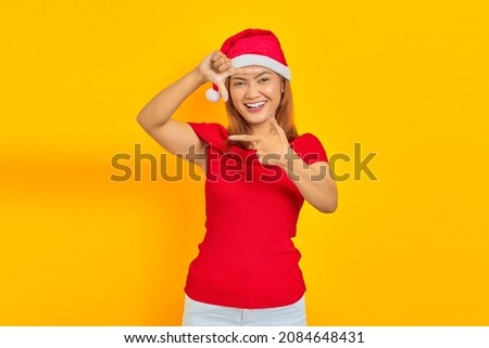 Portrait of cheerful young Asian woman wearing Christmas hat while making frame with fingers isolated on yellow background