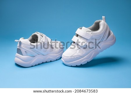 front view white child sneakers with velcro fasteners, one sneaker flying in the air and the other standing on the floor isolated on a blue background.