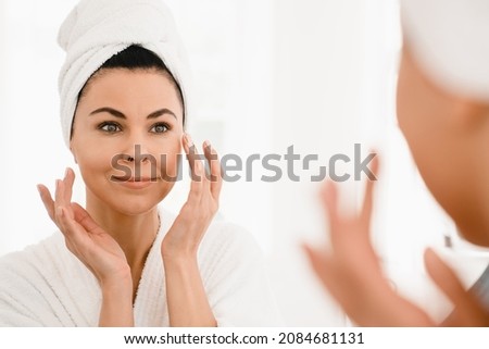 Good-looking caucasian middle-aged woman in turban and spa bathrobe after taking shower looking at the mirror while applying beauty creme moisturizer for anti-age anti-wrinkle effect Royalty-Free Stock Photo #2084681131