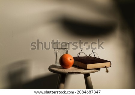 Still life of orange fruit, glass of water, a book and glasses on white wooden stool against white wall with sunlight and shadow
