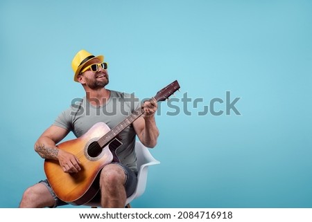 Handsome bearded guitarist in yellow hat and sunglasses sitting on chair and singing while playing on acoustic guitar isolated on blue background.