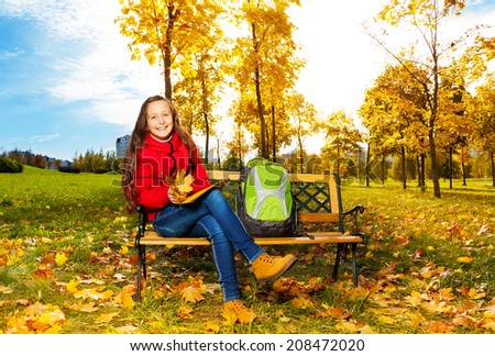 Autumn portrait of a 11 years old girl sitting on the bench in the park after school with backpack