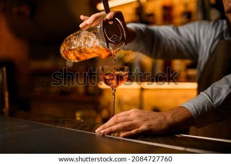 great view of a transparent mixing cup with cold cocktail with a strainer from which beverage is poured into a wine glass on the bar Royalty-Free Stock Photo #2084727760