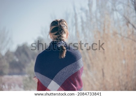 Detail of the girl's back with her hair collected in a braid looking and appreciating the snowy landscape on a sunny day with a with a scarf around her body. 
