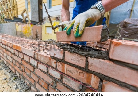Bricklayer is near brickwork on exterior wall with putty knife in construction site. Royalty-Free Stock Photo #2084740414