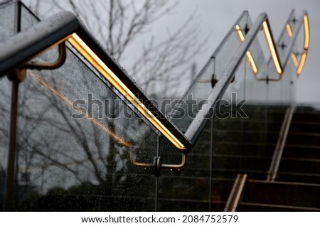 from the bottom of the handle is a recessed LED strip with a yellow light. the side of the stair railing is made of glued glass panels without frames. stainless steel railing bars on metal handles Royalty-Free Stock Photo #2084752579