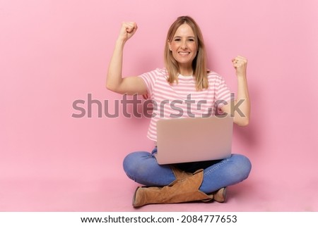 Young smiling woman in striped t-shirt using laptop computer making winner gesture sitting on floor over pink background.