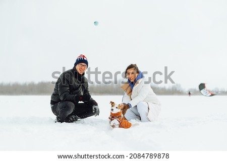 Family man and woman playing with a cute dog in a jack russell in the snow with the help of a ball with a smile on his face. Winter games in the snow on a walk with a pet.