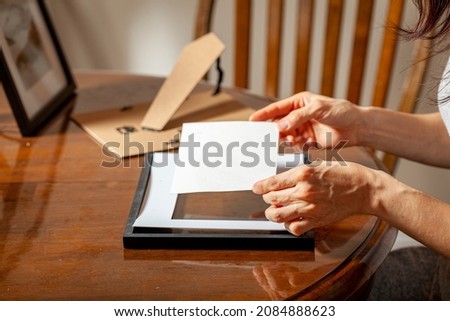 A young caucasian woman is placing a printed photograph into a picture frame with kickstand. There are other frames on the table. Tabletop picture framing concept.