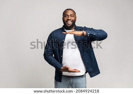 Portrait of smiling young handsome African American guy in blue shirt standing isolated over gray background and looking at the camera, showing measuring size with hands, copy space mockup image