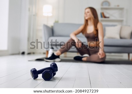 Blurred image of young beautiful athletic girl in leggings and top makes an exercises at home. Healthy lifestyle. In the foreground dumbbells. Royalty-Free Stock Photo #2084927239