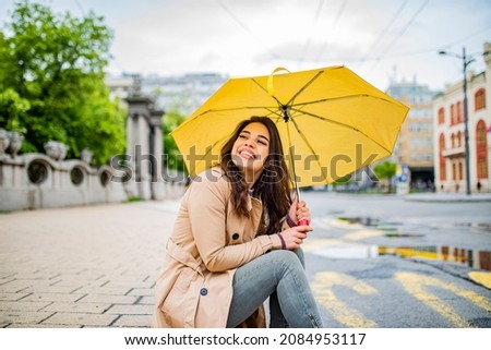 Good mood at any weather. Attractive young smiling woman carrying umbrella and adjusting her coat while walking by the street. The young woman smiles and laughs under the rain