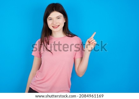 Caucasian woman wearing pink T-shirt over blue background looking at camera indicating finger empty space sales