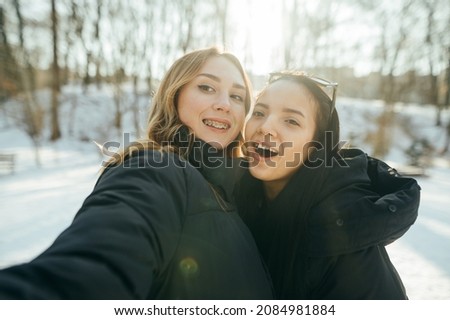 Two happy girlfriends in warm clothes take a selfie while walking in a snowy forest in sunny weather, look into the camera and smile.