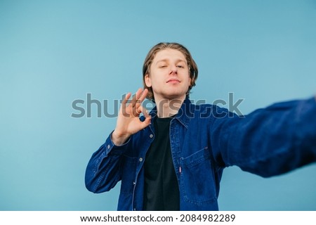 Handsome young man in a blue shirt takes a selfie on a blue background and shows a gesture ok with a smile on his face. Isolated.