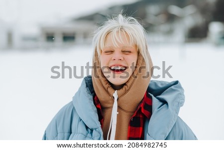 Close portrait of happy teen girl with blond hair on a snowy snowy background in winter, smiling with closed eyes.