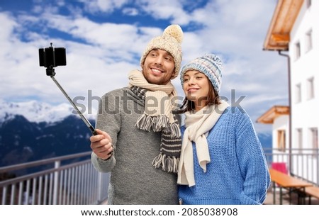 travel, tourism and winter holidays concept - happy couple in knitted hats and scarves taking picture by smartphone on selfie stick over mountains and ski resort background