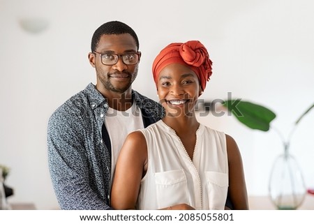 Portrait of cheerful middle aged couple embracing at home. Romantic mid adult black man hugging beautiful wife with traditional red turban while smiling and looking at camera. Mature african couple. Royalty-Free Stock Photo #2085055801