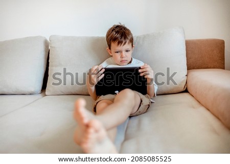 Little boy is lying on his bed in the morning, using a digital tablet. Shot of a young boy using tablet while lying on couch at home. Portrait of a young child at home watching cartoon on the tablet.