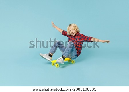 Full length of cheerful little curly kid boy 10s in basic red checkered shirt sitting on skateboard spreading hands isolated on blue background children studio portrait. Childhood lifestyle concept