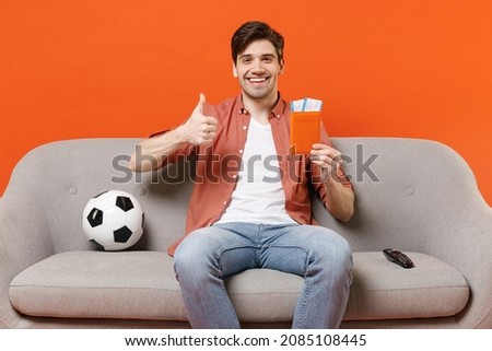 Traveler tourist man football fan in shirt sit sofa near ball home watch tv hold passport ticket show thumb up isolated on orange background Passenger travel abroad weekend getaway Air flight concept Royalty-Free Stock Photo #2085108445