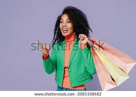 Young black curly woman 20s wears green shirt hold package bags with purchases on shoulder point on workspace area copy space mock up isolated on plain pastel light violet background studio portrait